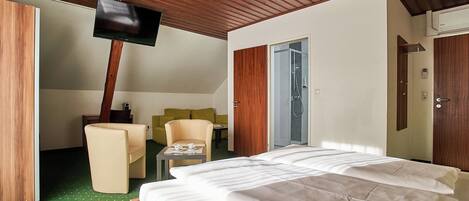 A hotel room with a bed, a television mounted on the wall, a small table with two cups, and a door leading to a shower area.