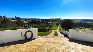 Ein gepflasterter Weg führt zu einer malerischen Aussicht auf eine grüne Landschaft mit blauem Himmel und strahlender Sonne.