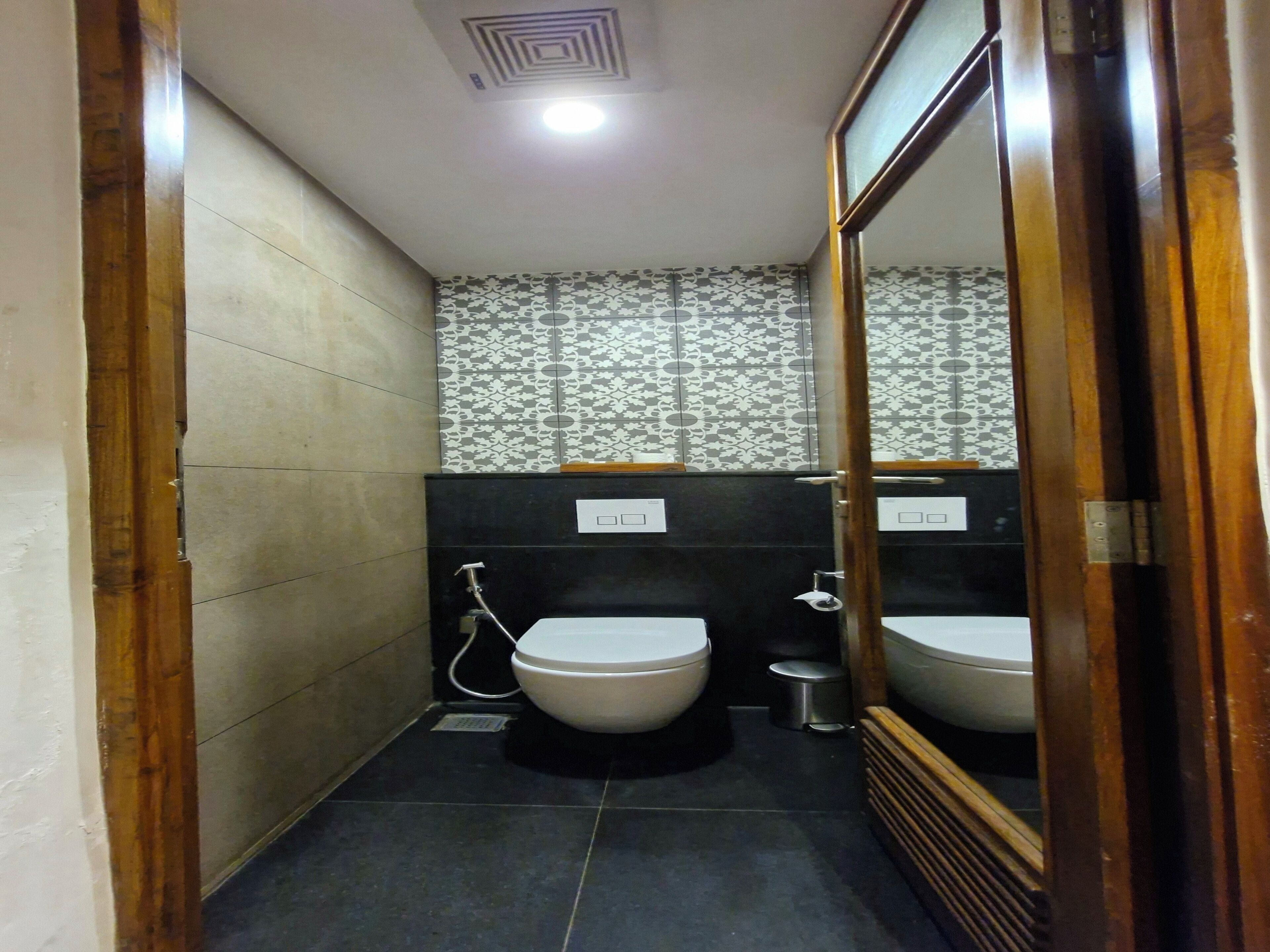 A small, narrow bathroom with two toilets, a sink, and a shower. The walls are tiled, and there is a patterned wallpaper above the sink.
