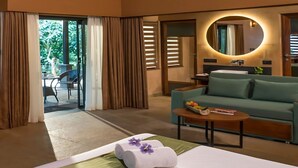 A hotel room with a bed, a green sofa, a small table with fruit, a round mirror, and a view of the outdoors. - Klov Resort & Spa (Mananthavady)
