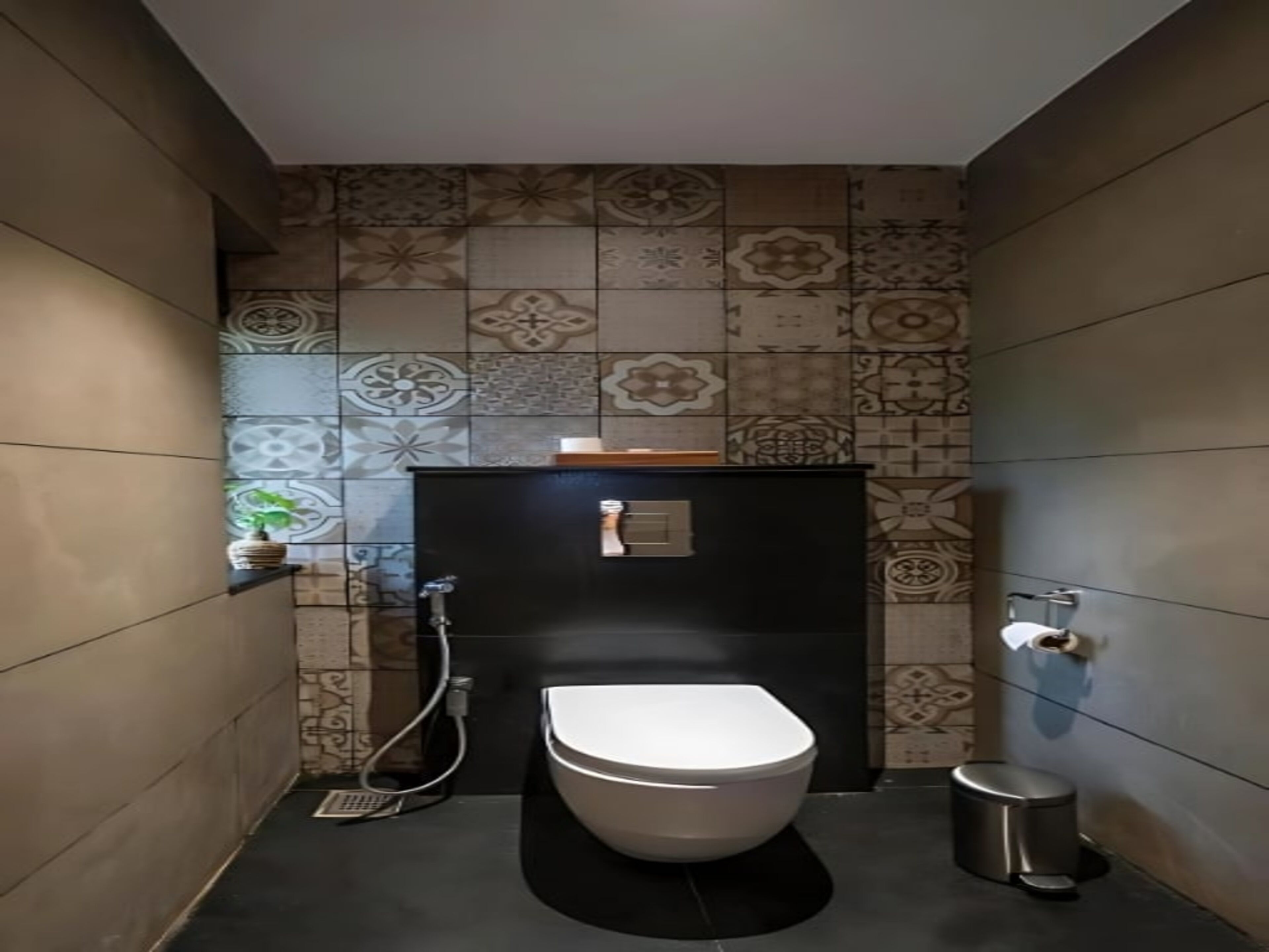 A modern bathroom with a white toilet, a shower, and a trash bin. The walls have patterned tiles, and there's a small shelf with a plant.