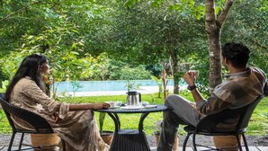 Two people seated at an outdoor table with a coffee pot, cups, and a plate, surrounded by greenery and a blue wall in the background. - Klov Resort & Spa (Mananthavady)
