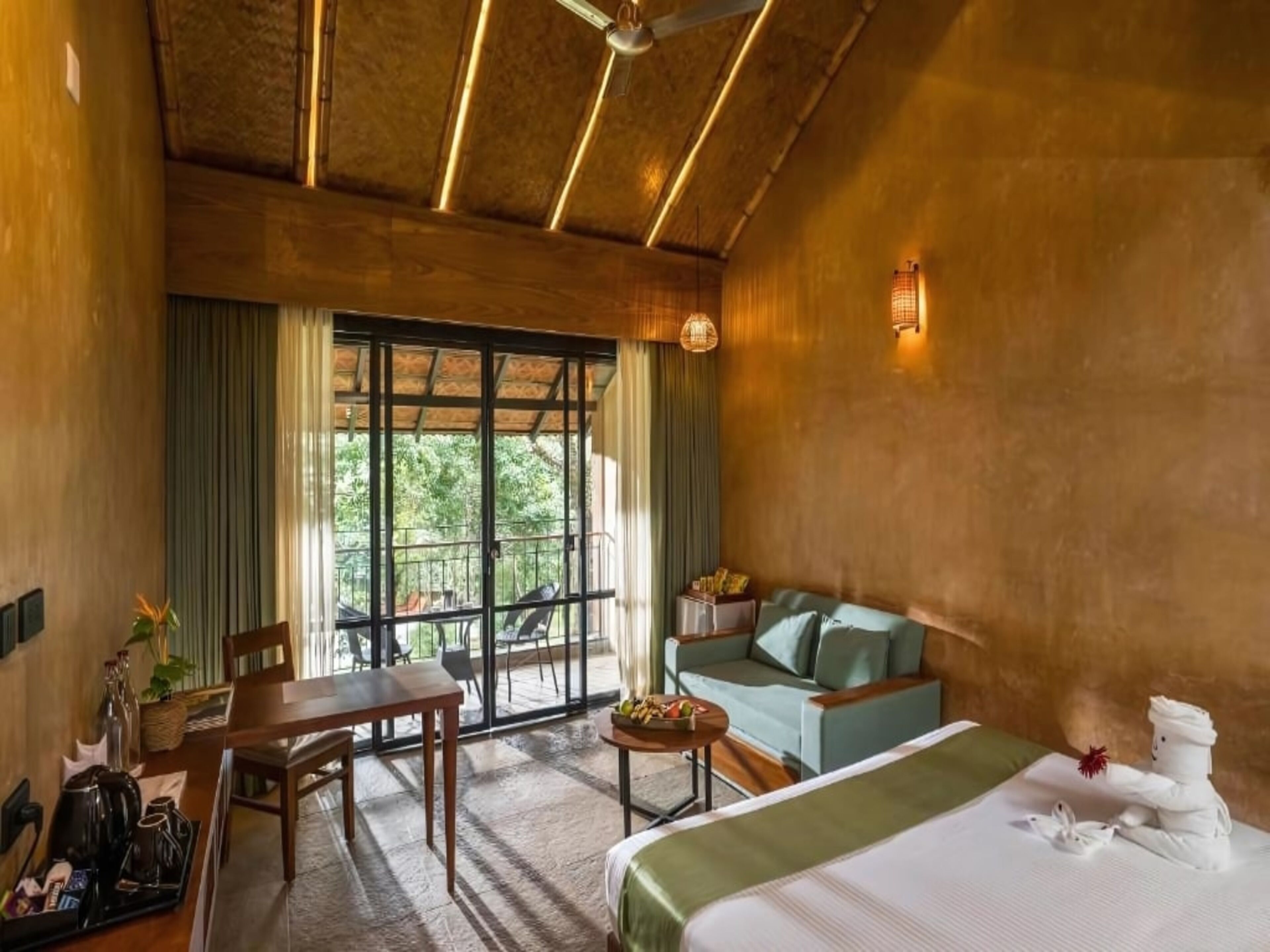 A hotel room with a bed, a sofa, a dining table, and a balcony with a view of trees.