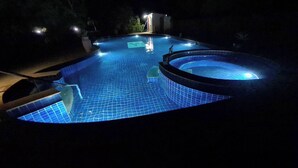 A swimming pool at night with tiled flooring and a hot tub area.