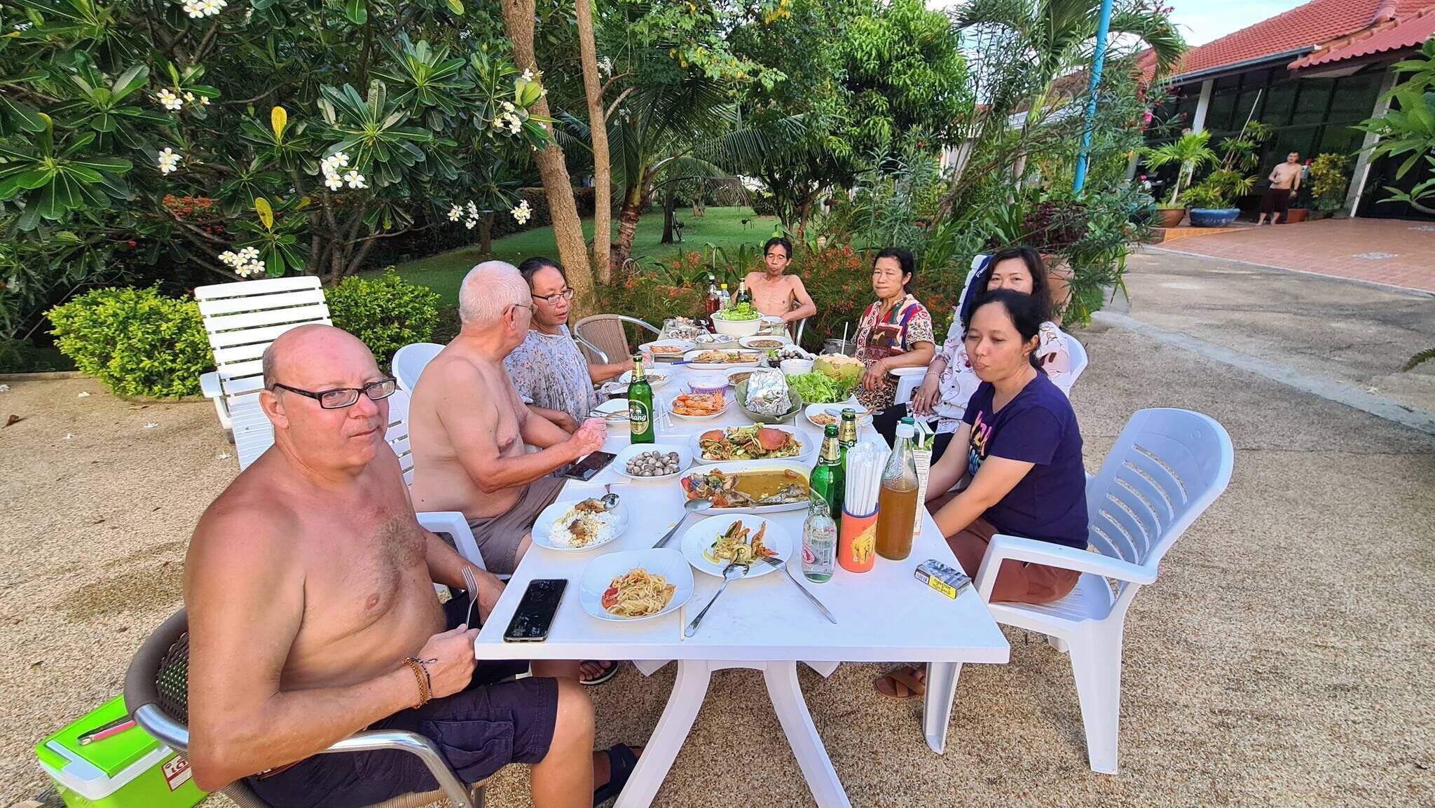 Un grupo de personas sentadas en una mesa al aire libre, con varios platos y bebidas, rodeadas de follaje.