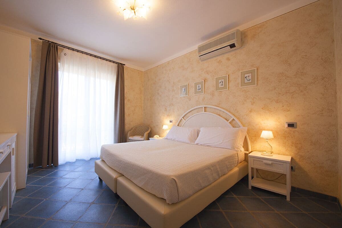 Un dormitorio con cama, mesitas de noche, una silla y un ventanal con cortinas.