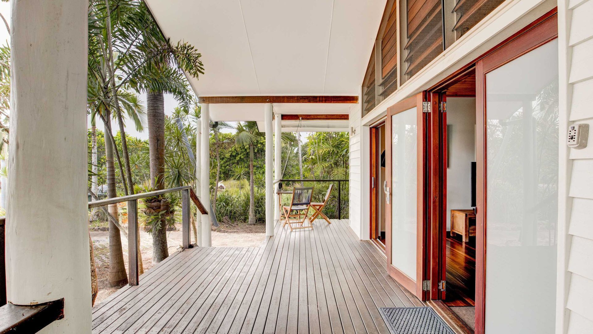 Una terraza de madera con una puerta de vidrio que da a una habitación, rodeada de vegetación tropical.