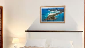 A bed with white linens, a nightstand with a lamp, and a framed coastal landscape painting on the wall.