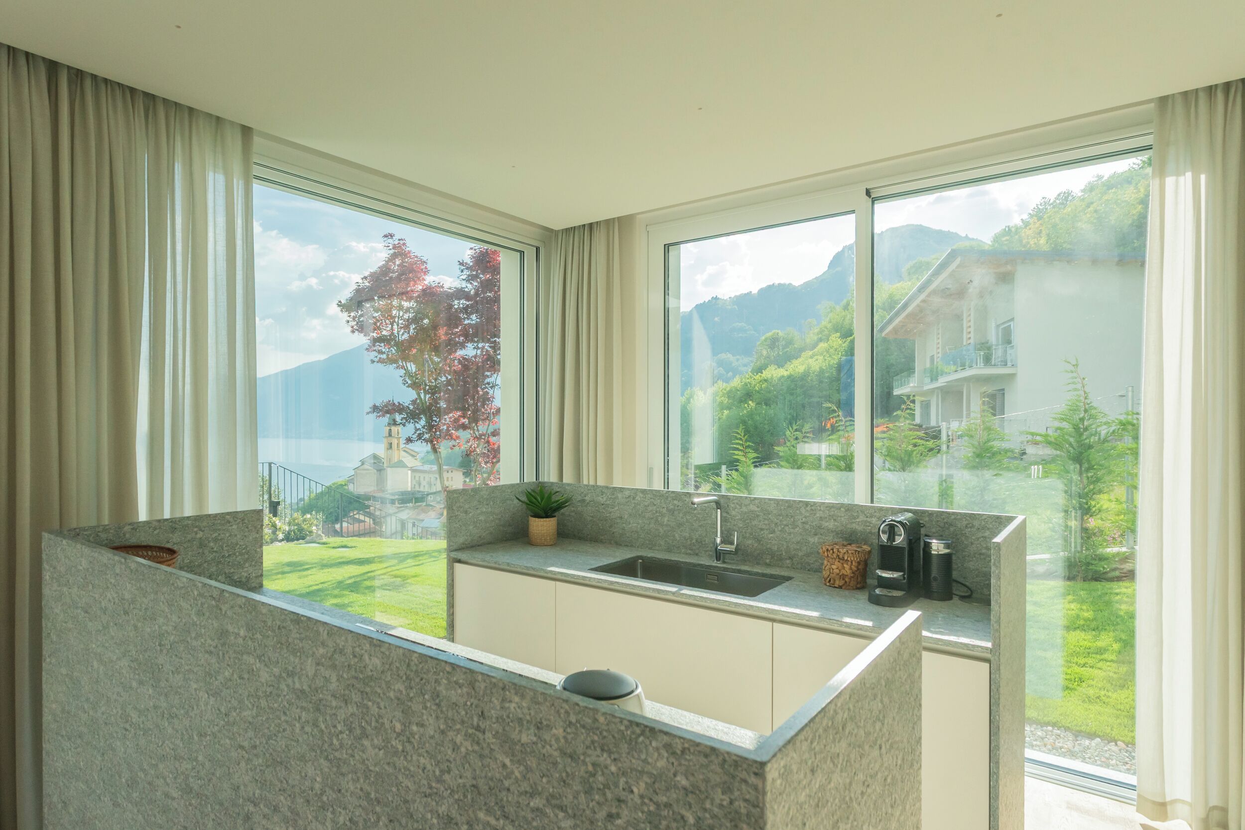 Una cocina moderna con encimera de granito, un lavabo y una cafetera. Cuenta con ventanas amplias con vistas a un paisaje verde y una montaña.