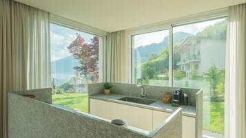 Una cocina moderna con encimera de granito, un lavabo y una cafetera. Cuenta con ventanas amplias con vistas a un paisaje verde y una montaña.