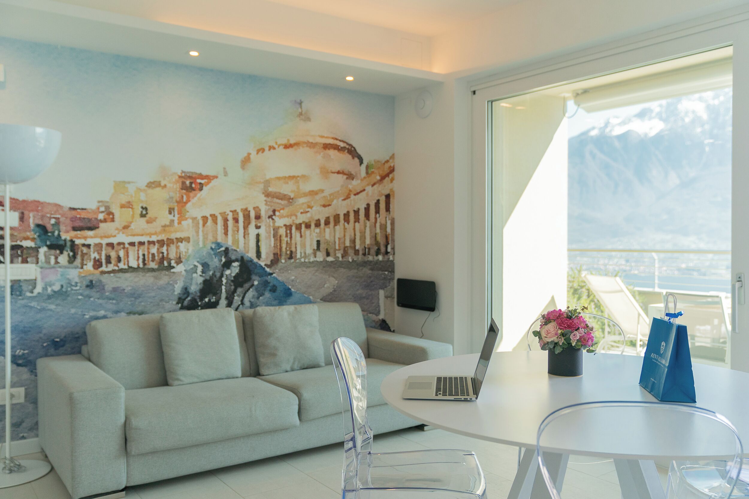 Un salon moderne avec un canapé, un fauteuil transparent, un ordinateur portable posé sur une table et une vue sur les montagnes par une grande fenêtre.