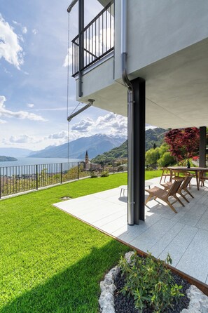 A terrace with a view of a lake and mountains, a dining area with chairs and a table, and a railing.