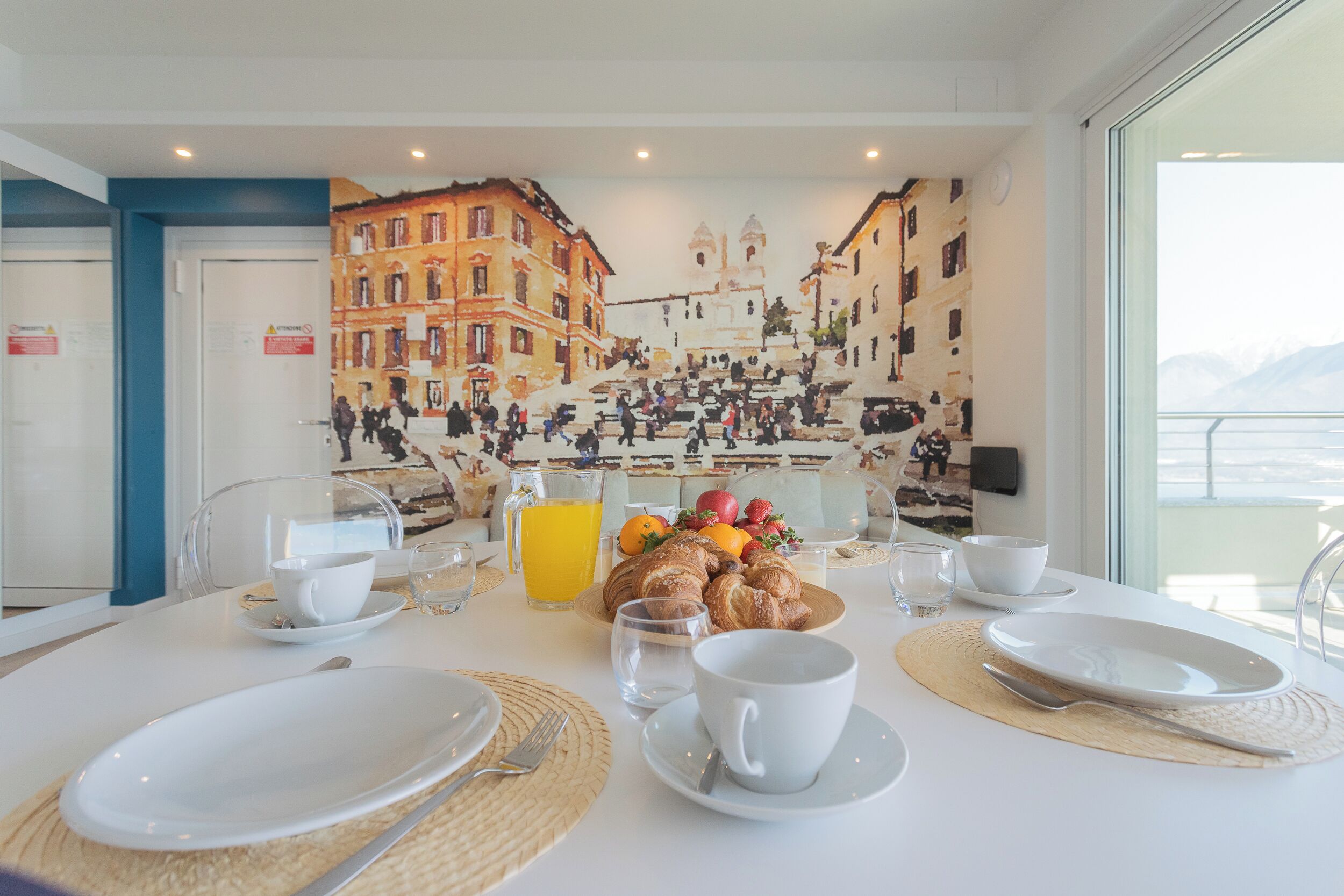 A breakfast table set with plates, cups, and croissants, with a mural of a bustling street scene in the background.