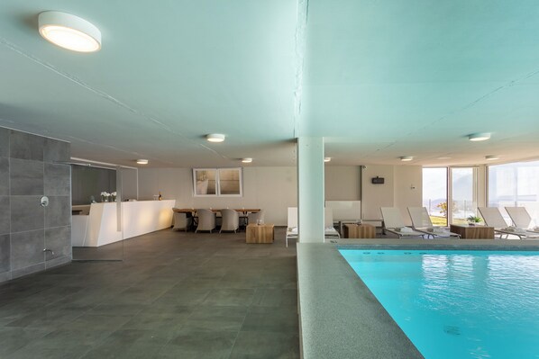 Un área de piscina interior moderna con vista despejada, sillas de descanso y una recepción.