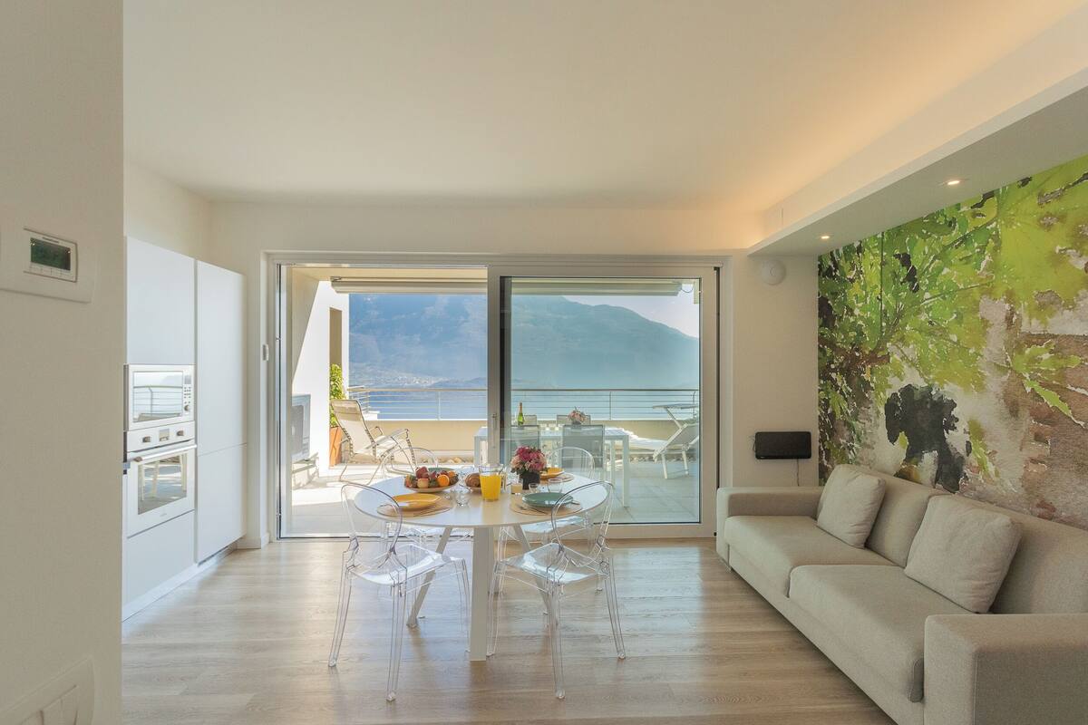 Uma sala de estar moderna com um sofá, uma mesa de jantar com cadeiras e vista para o mar através de grandes portas de vidro.