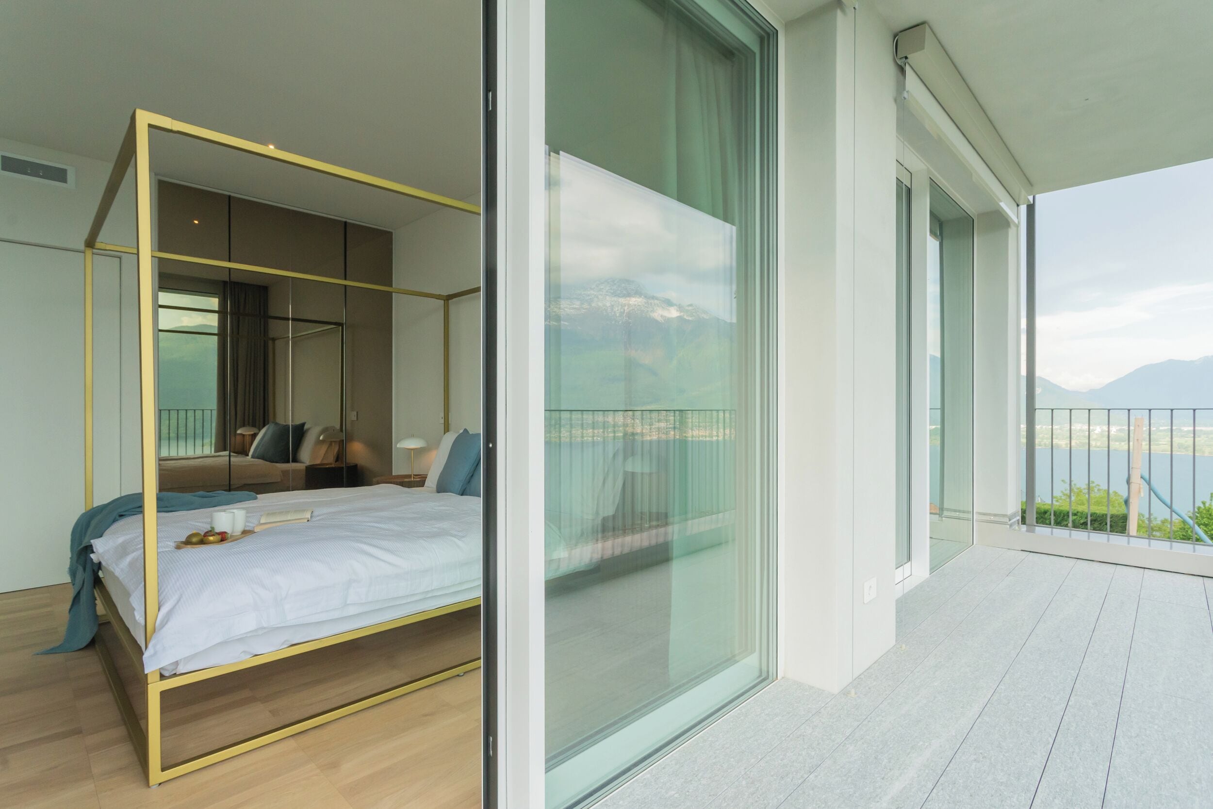 Une chambre moderne avec un lit à baldaquin, un balcon doté d’une rambarde en verre et une vue sur les montagnes.