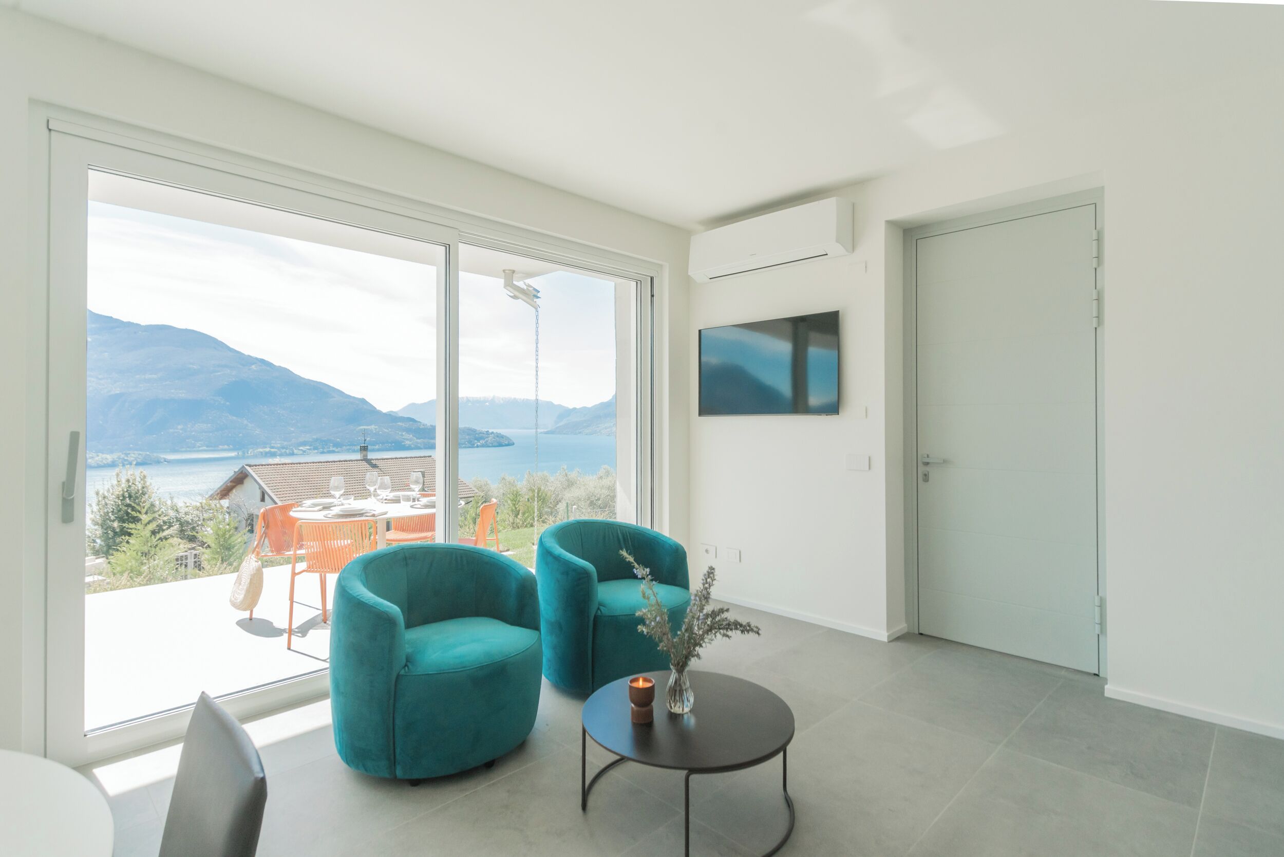 Een moderne kamer met een grote schuifdeur, twee teal fauteuils, een tafeltje met een vaas en een flatscreen televisie aan de muur.