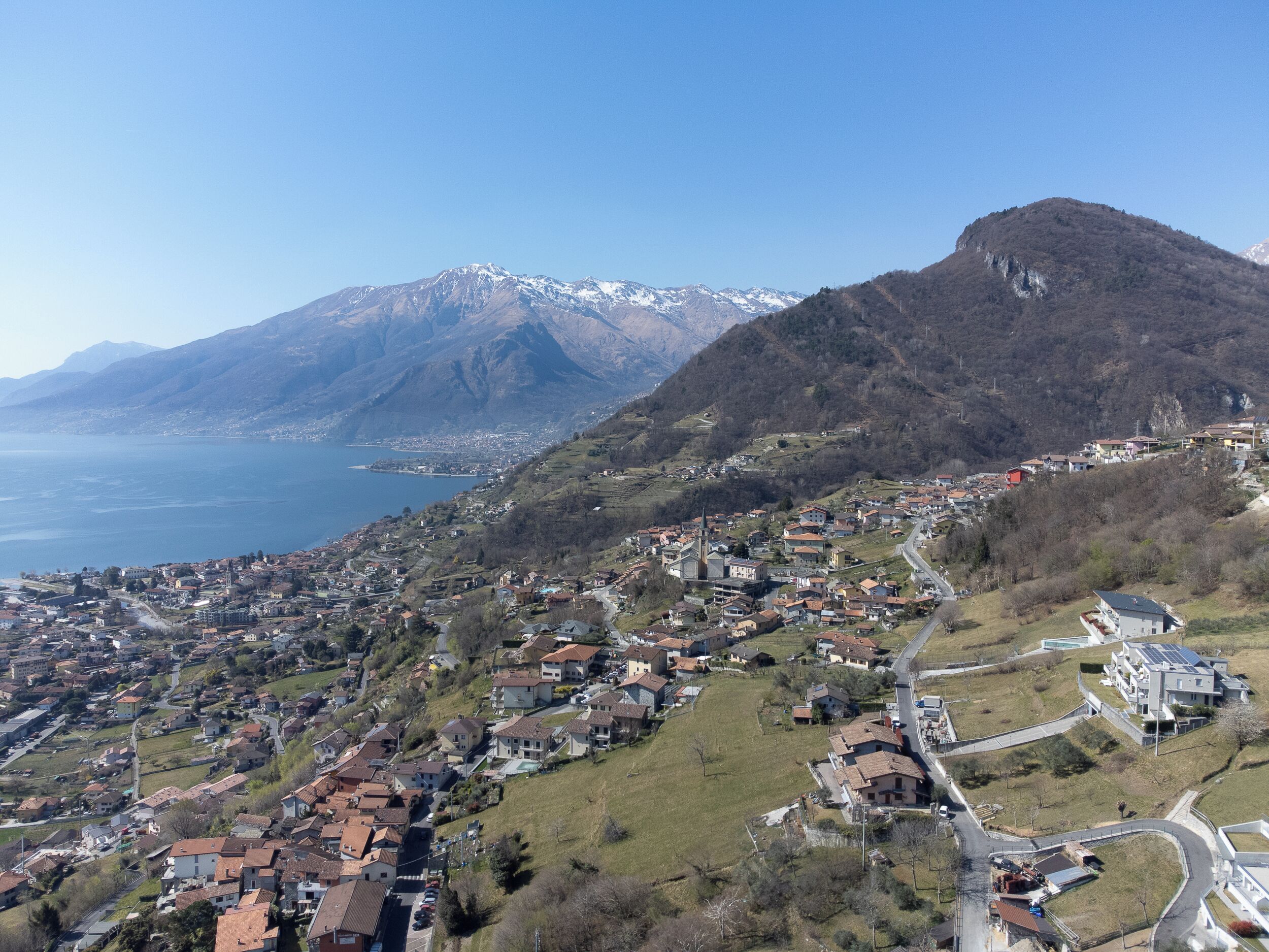 Eine Stadt in den Bergen, mit einer Mischung aus Wohn- und Geschäftsgebäuden, einer markanten Kirche und Blick auf See und Berge im Hintergrund.