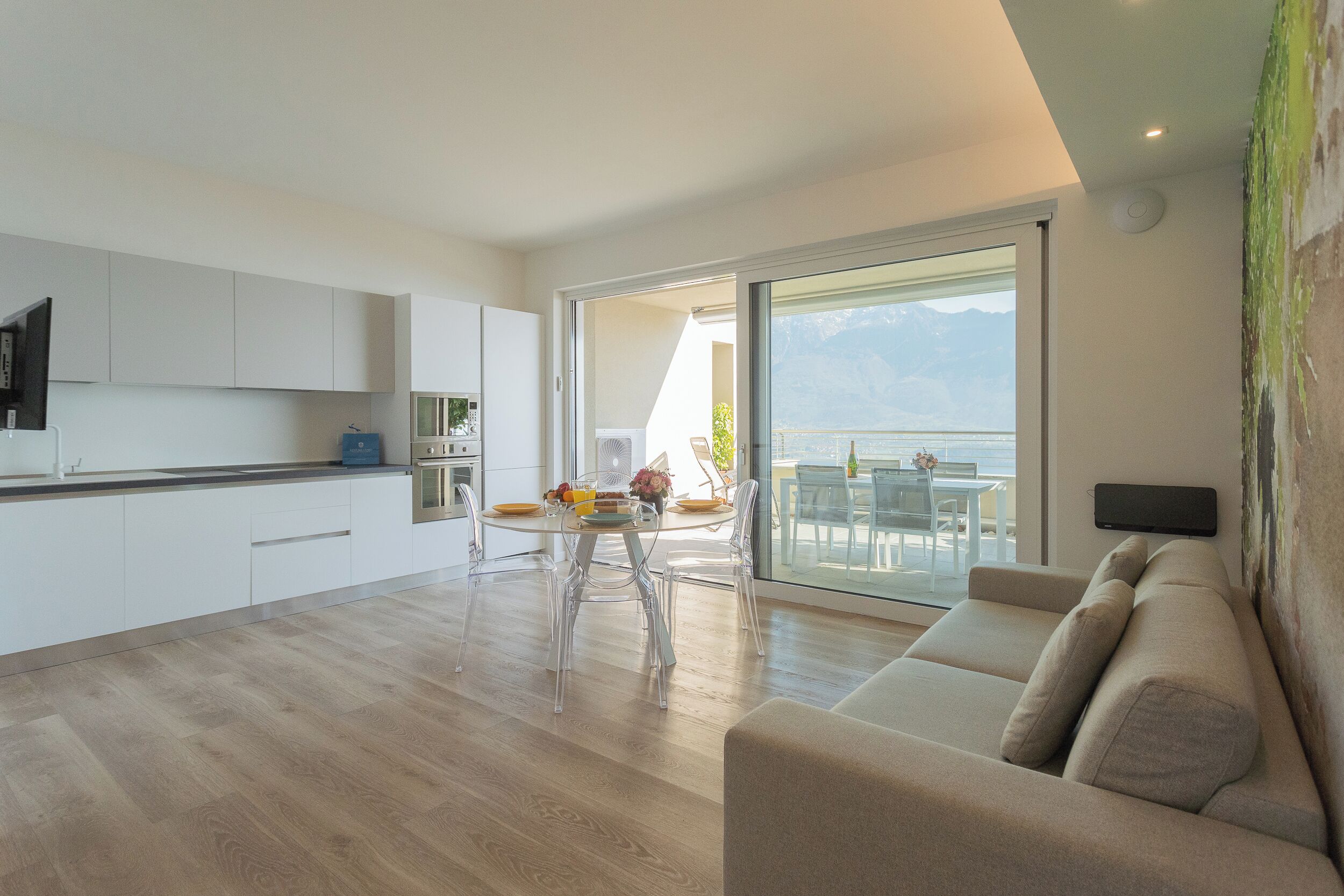 A modern kitchen with white cabinets, a dining area with a glass table and chairs, and a sofa with a view of the outdoors.