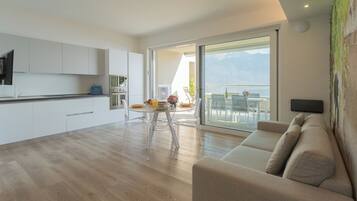 A modern kitchen with white cabinets, a dining area with a glass table and chairs, and a sofa with a view of the outdoors.