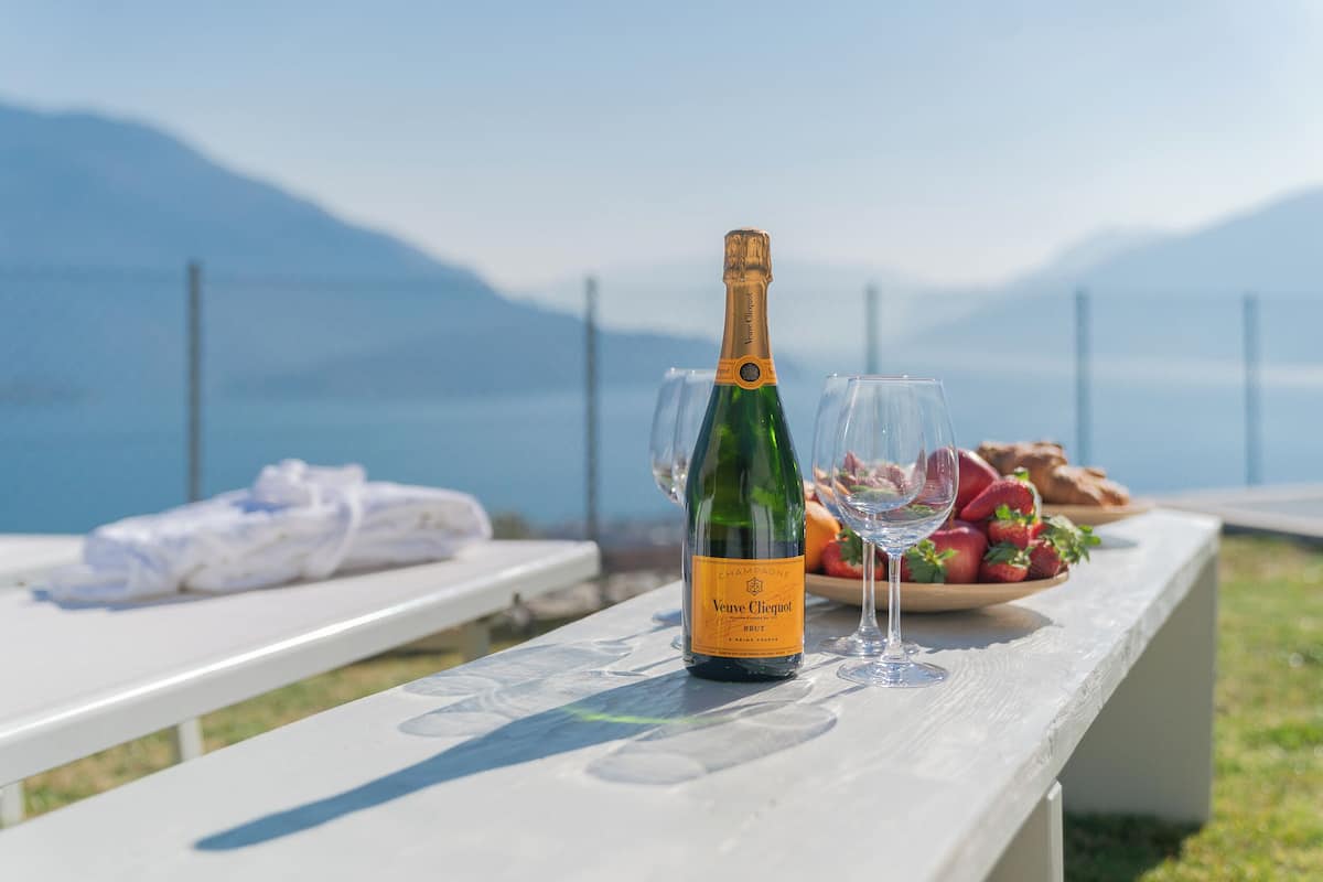 Uma garrafa de champanhe Veuve Clicquot e uma taça sobre uma mesa branca, com uma vista panorâmica de montanhas ao fundo.