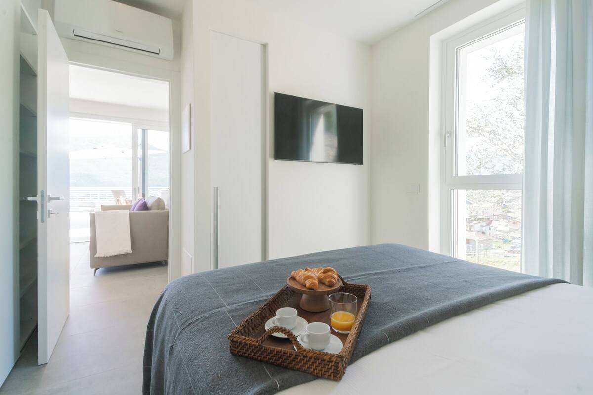 Quarto de hotel com cama, bandeja de café da manhã com croissants, xícaras e suco de laranja, televisão na parede e vista para o exterior.