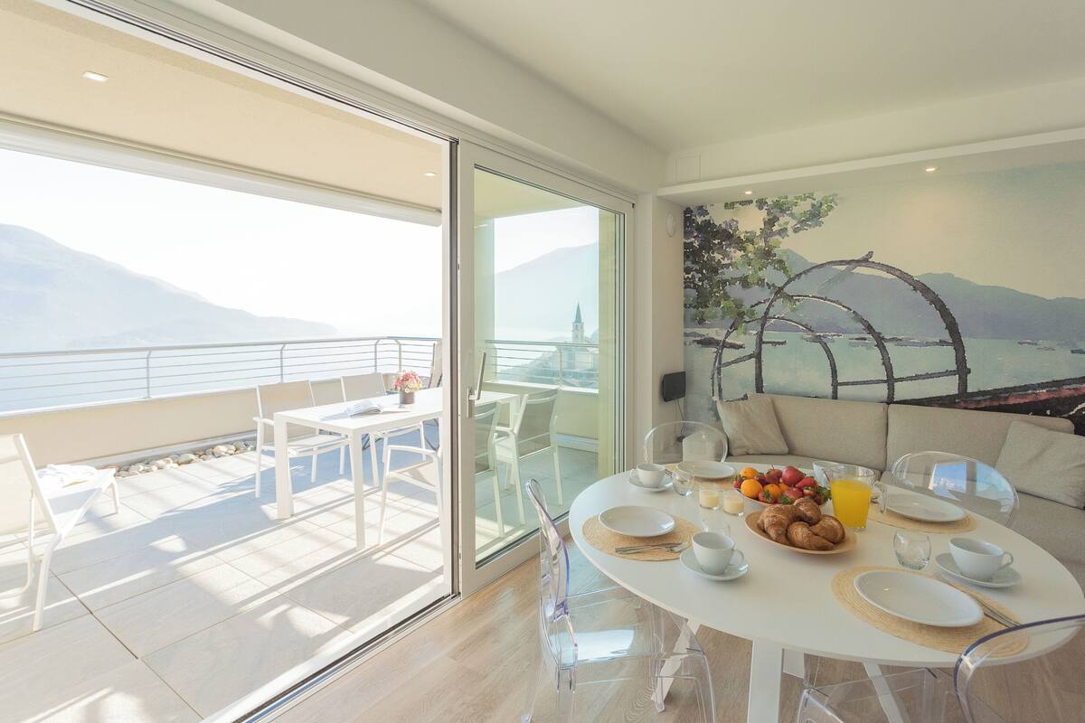 Uma área de refeições com uma mesa posta para o café da manhã, uma varanda com vista para montanhas e um quadro de uma paisagem na parede.