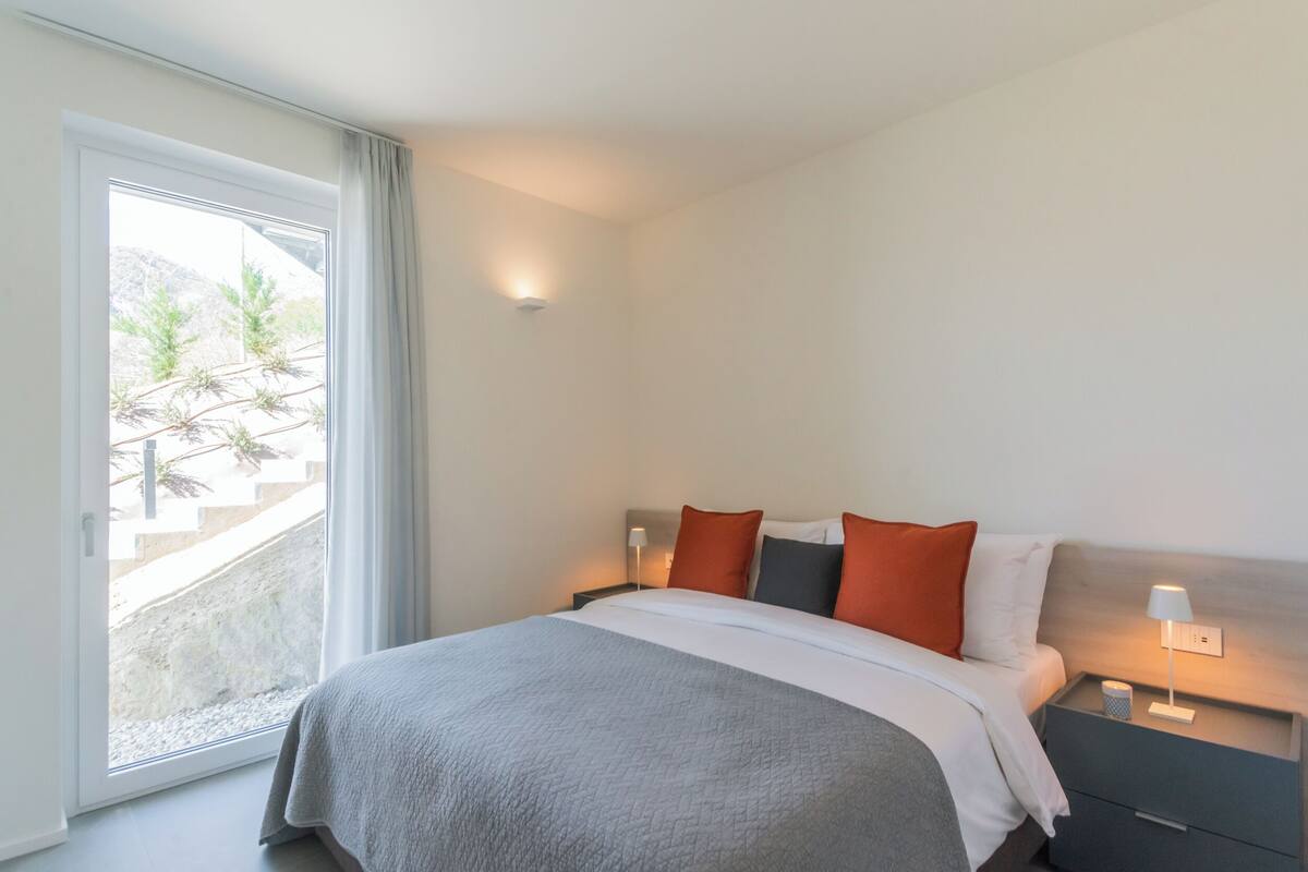 Quarto de hotel com cama, mesa de cabeceira, abajur e vista para uma varanda com plantas.