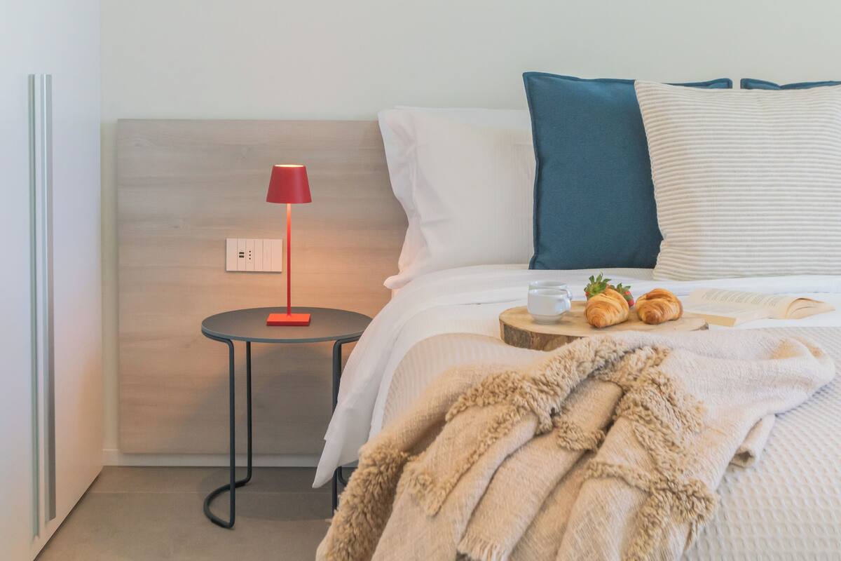 Quarto bem arrumado com uma cama, mesa de cabeceira com abajur e bandeja com croissants.