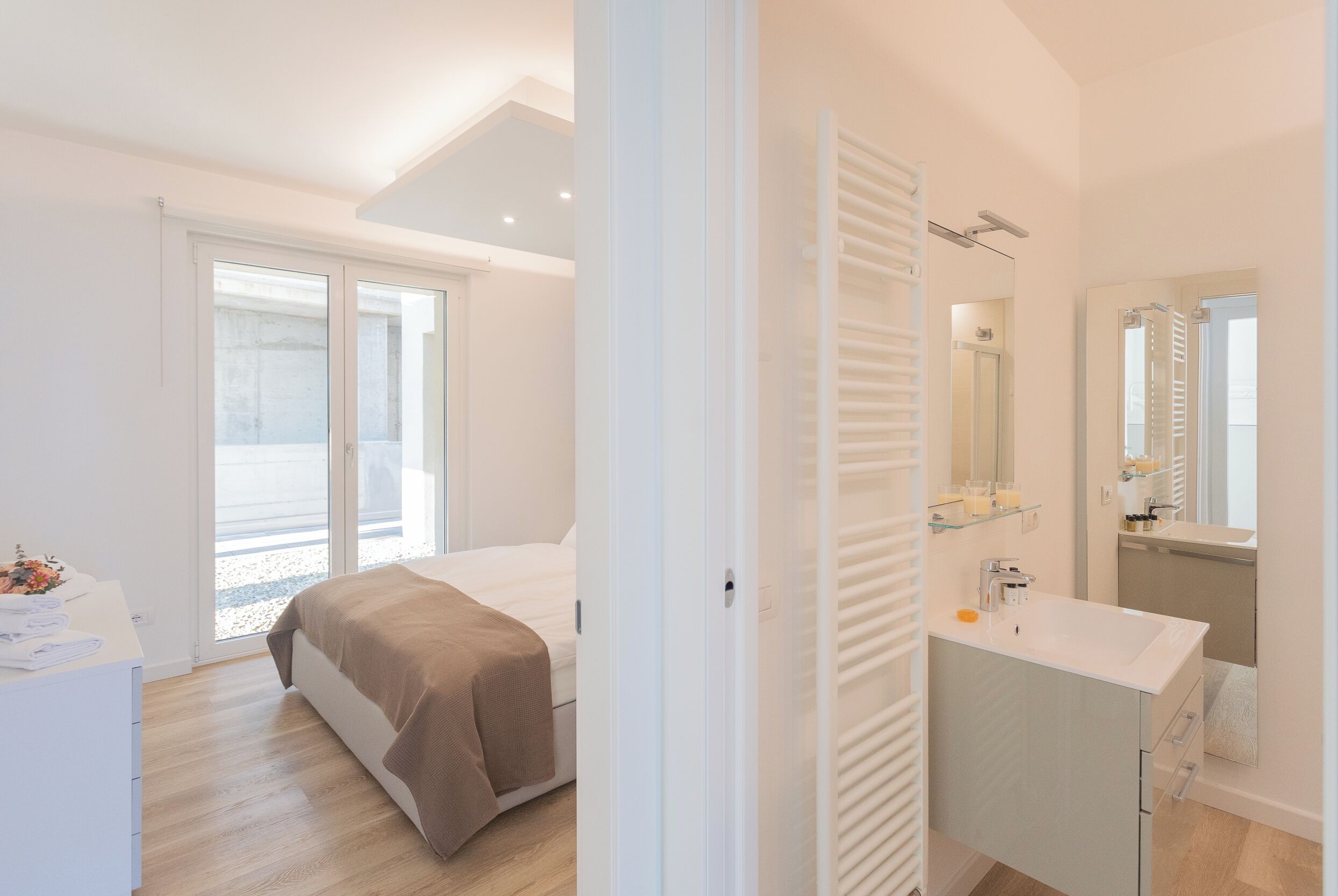 Une chambre à coucher moderne avec un lit, une table de chevet, une grande fenêtre et une salle de bain équipée d’un lavabo et d’un miroir.