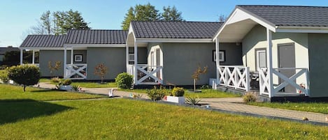 A row of modern, single-story mobile homes with white railings and grey roofs, surrounded by well-maintained grass and small shrubs.