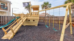 A wooden play structure with a slide, a small pirate-themed playhouse, and a swing set.