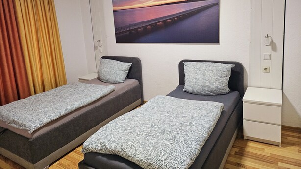 Une chambre avec deux lits simples, une petite table de chevet blanche, un tableau représentant un coucher de soleil sur un pont et une fenêtre avec des rideaux.