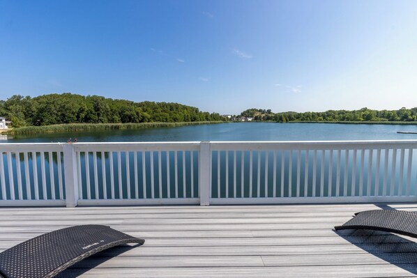 A wooden deck with white railings overlooking a calm lake, surrounded by trees and a distant hill.