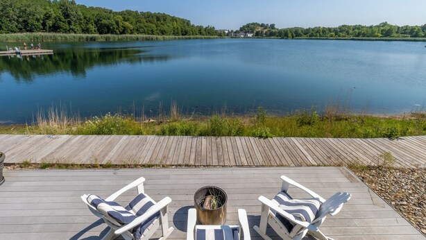 Eine Holzterrasse mit Liegestühlen, die auf einen ruhigen See blickt, umgeben von Grünpflanzen und einem klaren Himmel.