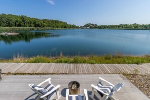 A wooden deck with lounge chairs facing a calm lake, surrounded by greenery and a clear sky.