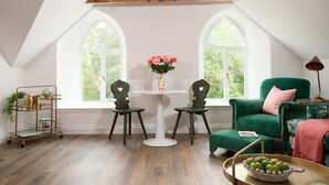 A room with a sloped ceiling, two large arched windows, a green armchair, a white table with chairs, and a small side table with a bowl of apples.