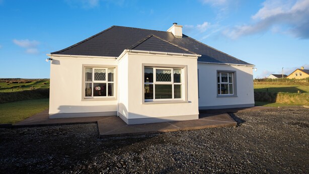 A single-story house with a dark roof and white exterior walls, featuring two windows with white frames and a small porch area.