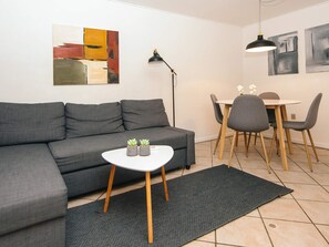 A modern living room with a grey sofa, a small white table with two potted plants, a dining area with a round table and chairs, and abstract art on the walls.