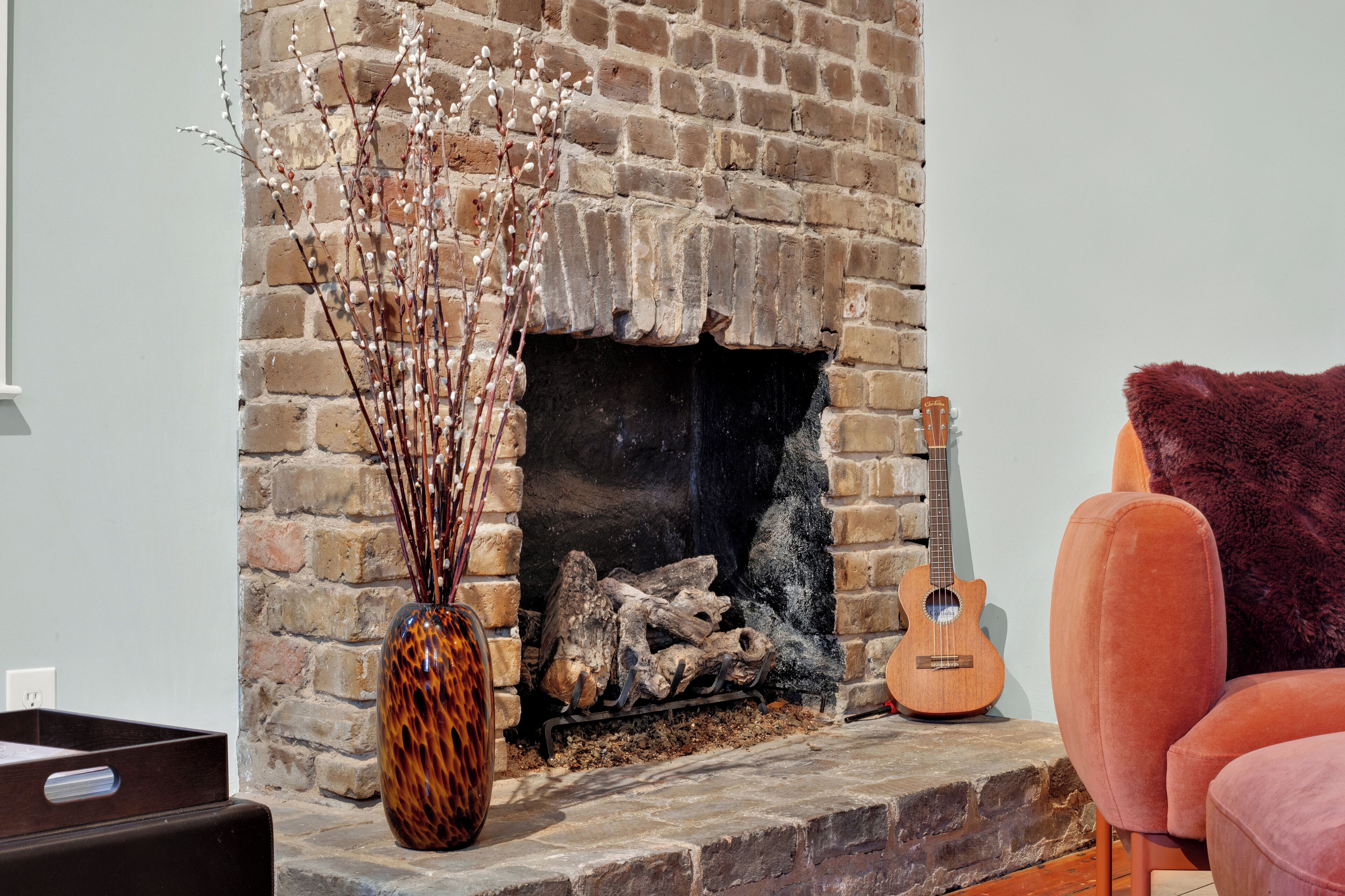 A fireplace with a brick wall, a guitar, and a vase with branches.