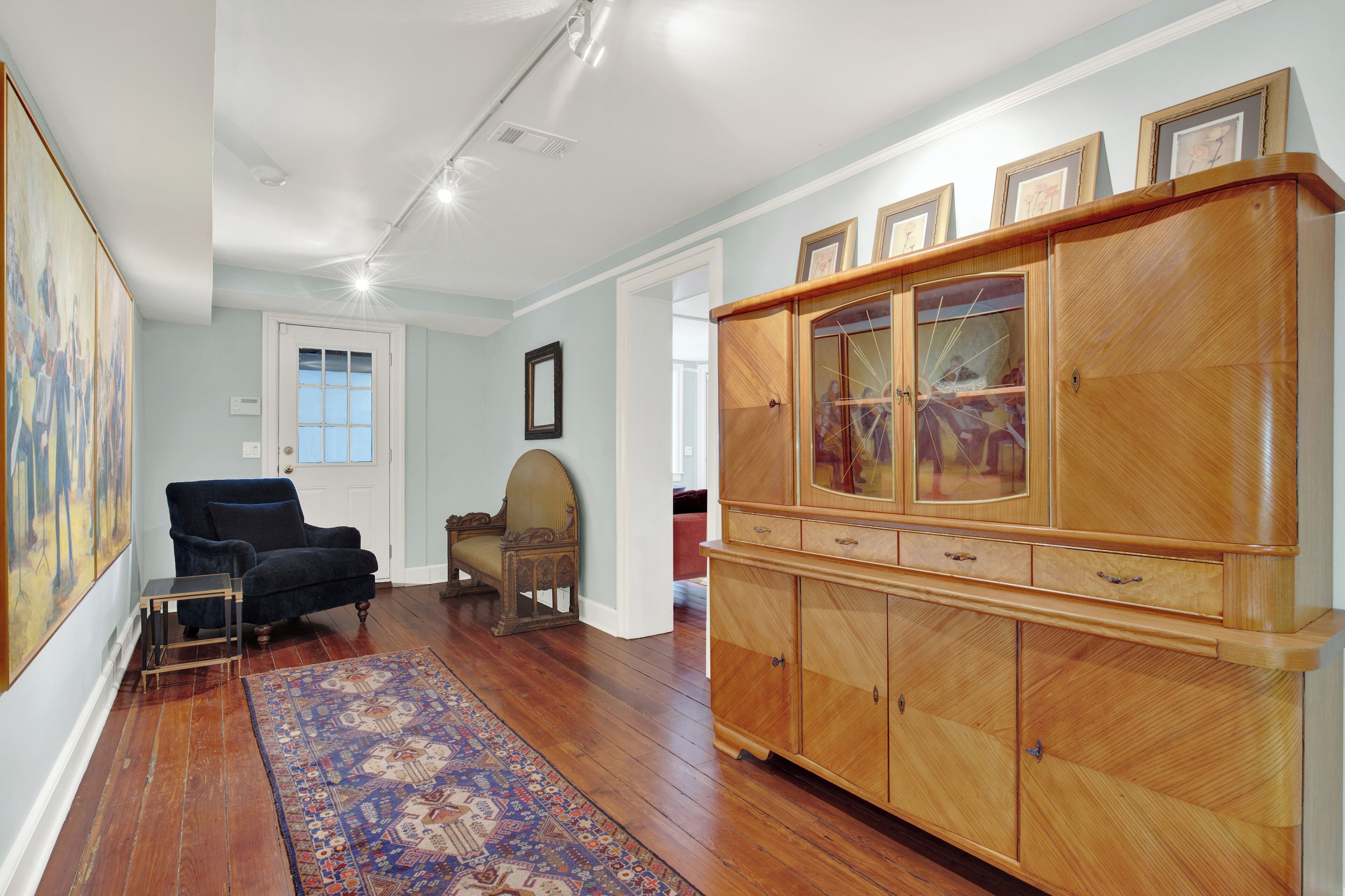 Une pièce spacieuse dotée d’un parquet, d’une grande armoire en bois aux portes vitrées, d’un fauteuil bleu et d’un tapis à motifs.