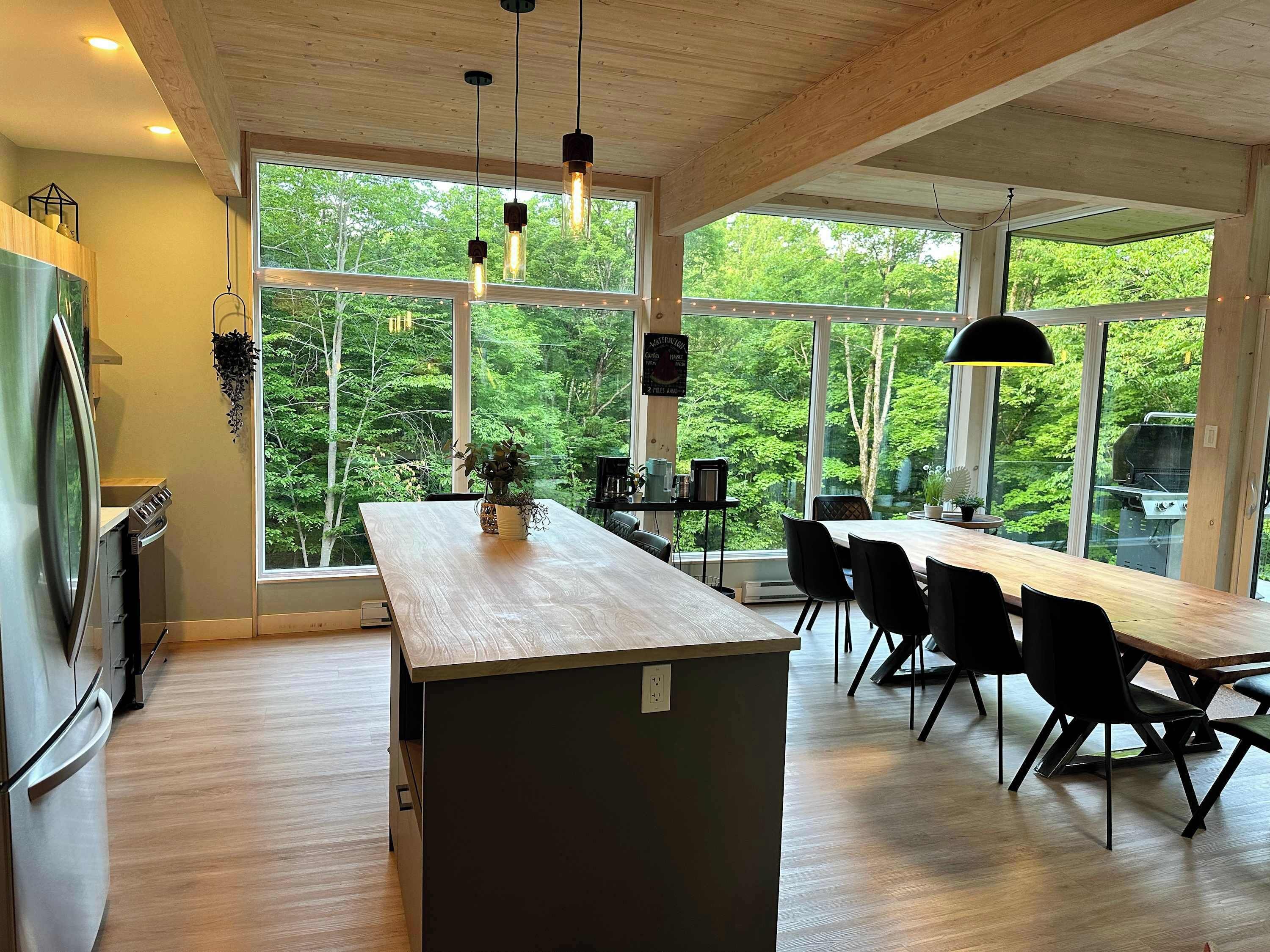 A modern kitchen with a large island, dining area with chairs, and large windows offering a view of the outdoors.