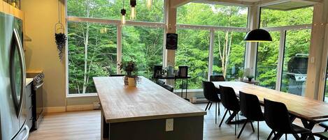 A modern kitchen with a large island, dining area with chairs, and large windows offering a view of the outdoors.