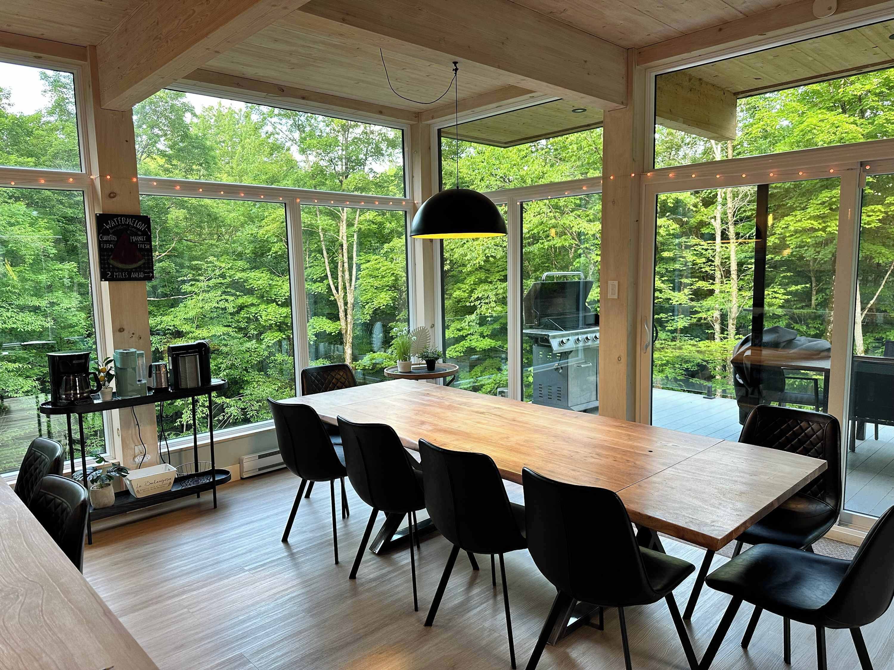 Un vaste espace dédié à la salle à manger, avec une grande table en bois et des chaises noires, le tout entouré de grandes fenêtres offrant une vue sur une zone boisée.