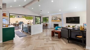 A modern kitchen with a central island, dining area with a sofa and table, and a TV mounted on the wall. - Maison Du Soleil – Luxury Palm Springs Retreat (Palm Springs)