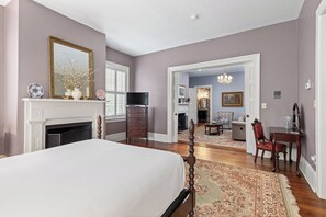 Une chambre avec un grand lit, une cheminée, une télévision et un couloir menant à une salle à manger.