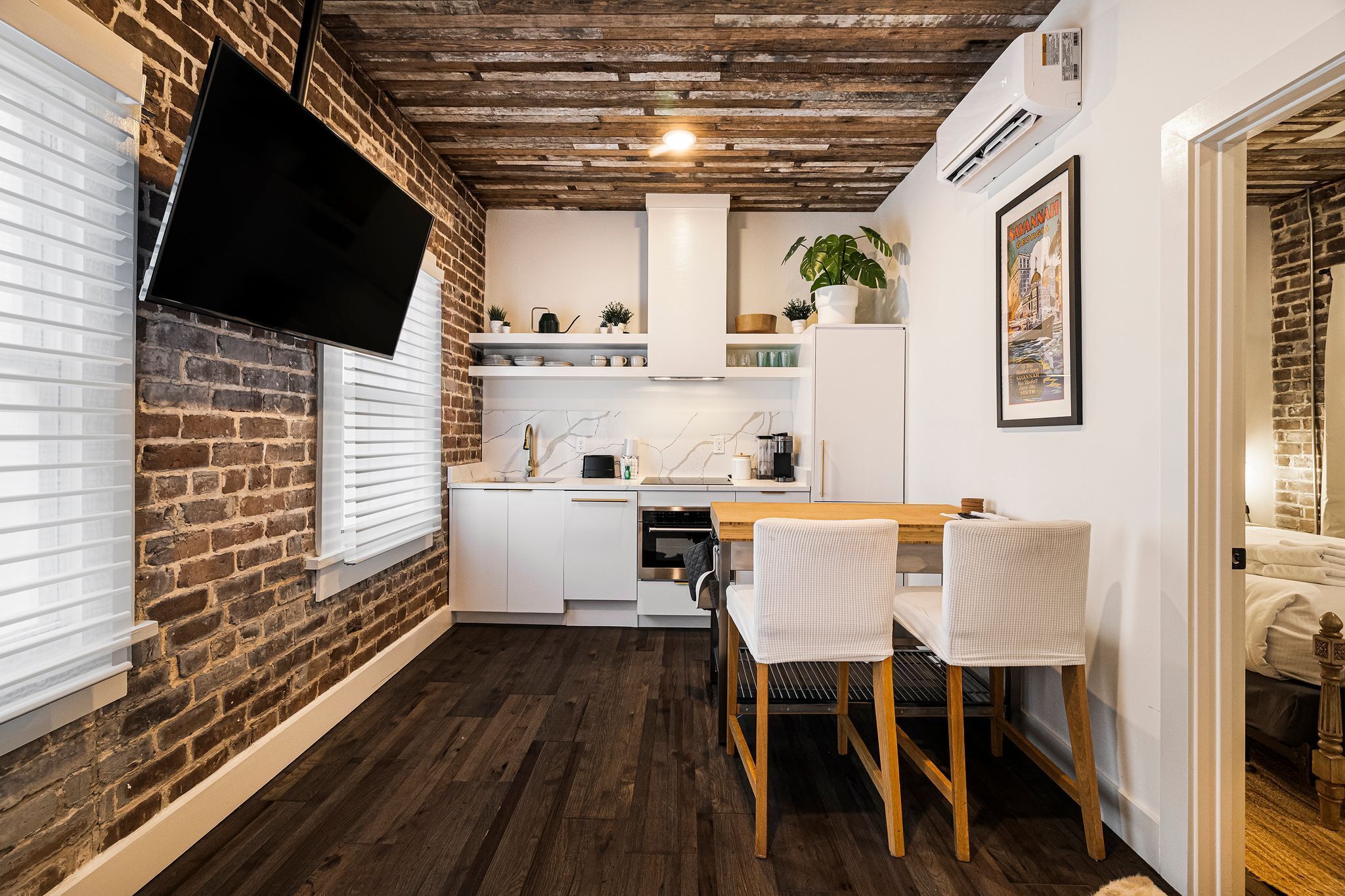 A compact kitchen with white cabinets, a wooden dining table, and a TV mounted on the brick wall.