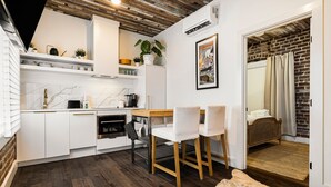 Eine moderne Küche mit weißen Schränken, einem Holztisch und Stühlen. Ein kleiner Essbereich mit Teppich und ein Bett, dessen Hintergrund eine Ziegelwand ist.