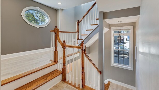 A staircase with wooden railings and white balusters, a round window with a view of palm trees, and a door with a view of parked cars.