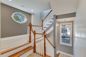 A staircase with wooden railings and white balusters, a round window with a view of palm trees, and a door with a view of parked cars.